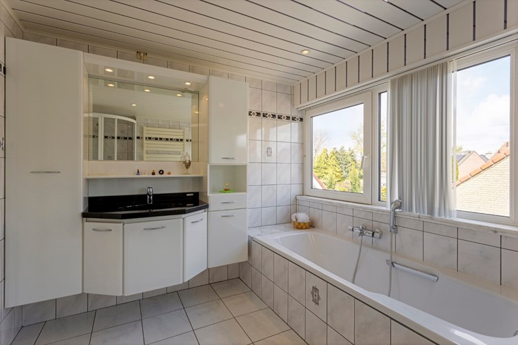 Ruime badkamer met een lichte tegelvloer, volledig licht betegelde wanden en een MDF plafond met inbouwspots. Met een kunststof ligbad met thermostaatkraan en een badmeubel met vaste wastafel. 