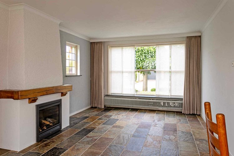 De woonkamer is voorzien van een leisteen tegelvloer met vloerverwarming. Veel daglicht via een groot hardhouten raamkozijn met roeden aan de voorzijde en een raamkozijn met glas-in-lood in de zijgevel.