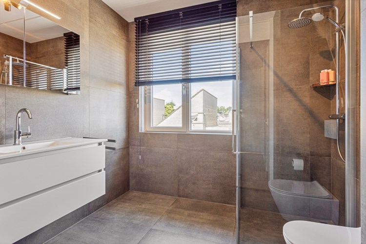 Moderne badkamer met volledig betegelde wanden en een spuitwerk plafond. Met een wandcloset met opzetplateau, een glazen douchecabine met thermostaatkraan en een spiegelkast met verlichting. Met mechanische afzuiging en een kunststof raamkozijn (draai-/kiep) met HR++ beglazing.