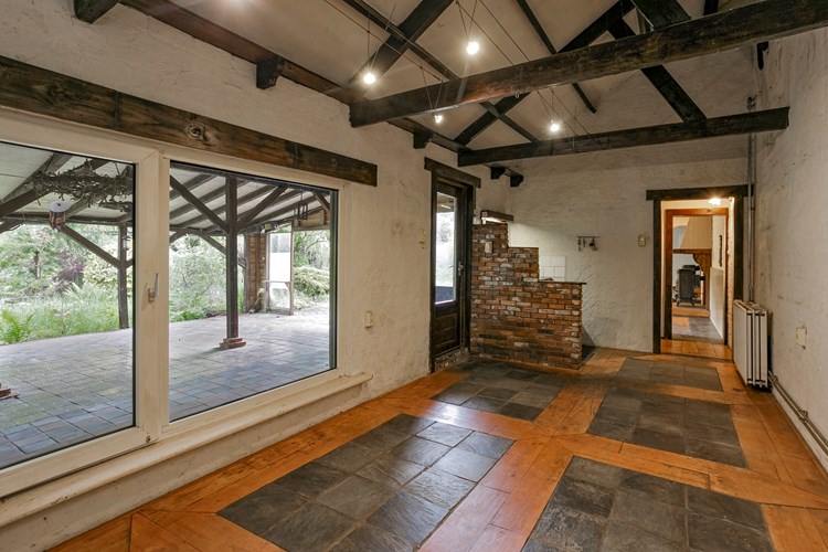 Sfeervolle tuinkamer met een planken- en tegelvloer, grof gestucte wanden en een hoog oplopend plafond. Achter het gemetselde muurtje bevindt zich het keukenblokje.