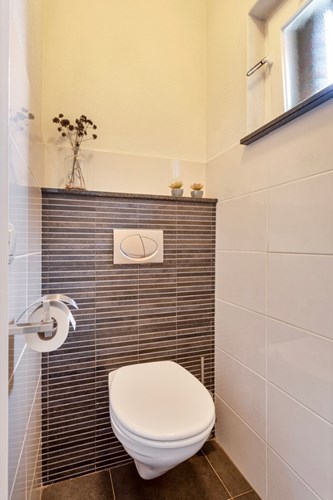 Modern toilet met een antraciet tegelvloer, gedeeltelijk betegelde wanden en een stucwerk plafond. Natuurlijke ventilatie middels een houten raamkozijn. 