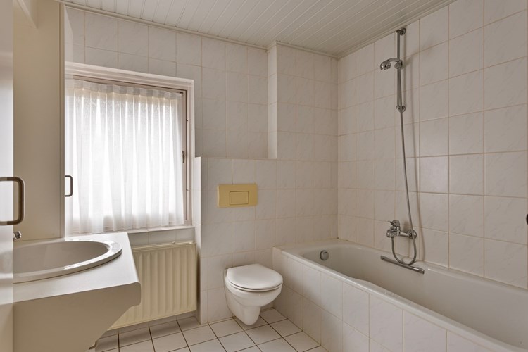 Badkamer met een lichte tegelvloer, volledig licht betegelde wanden en een schroten plafond. Met een wandcloset en een ligbad met douchemogelijkheid.