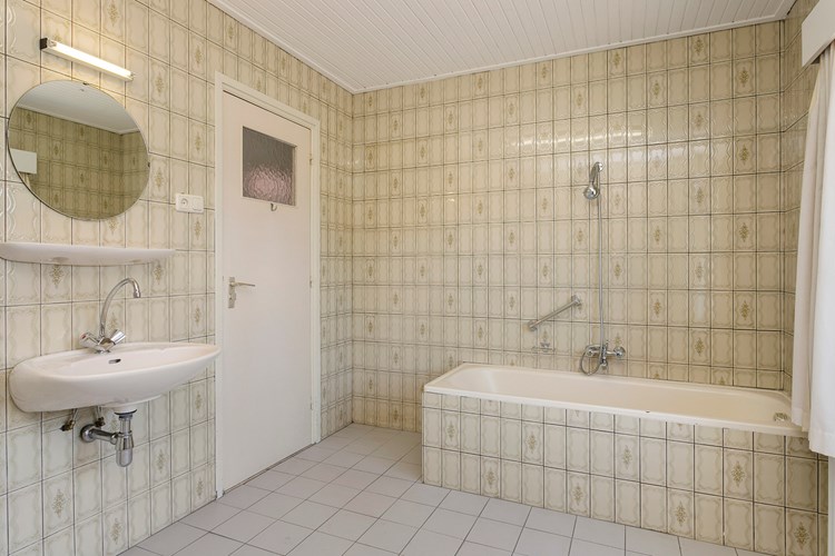 De ruime badkamer is volledig betegeld en heeft een schroten plafond. Met een stalen ligbad en een vaste wastafel met planchet, een spiegel en verlichting. 