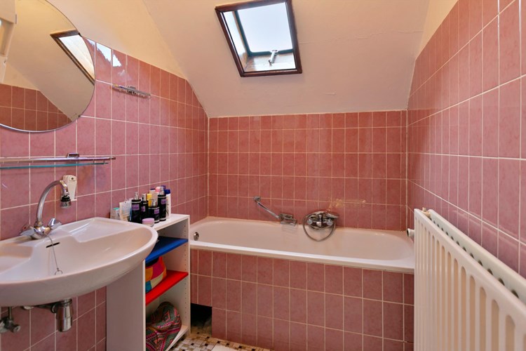 Badkamer met een tegelvloer, gedeeltelijk betegelde wanden en een stucwerk plafond. Met een ligbad en een wastafel met spiegel. Daglicht via een dakraampje. 