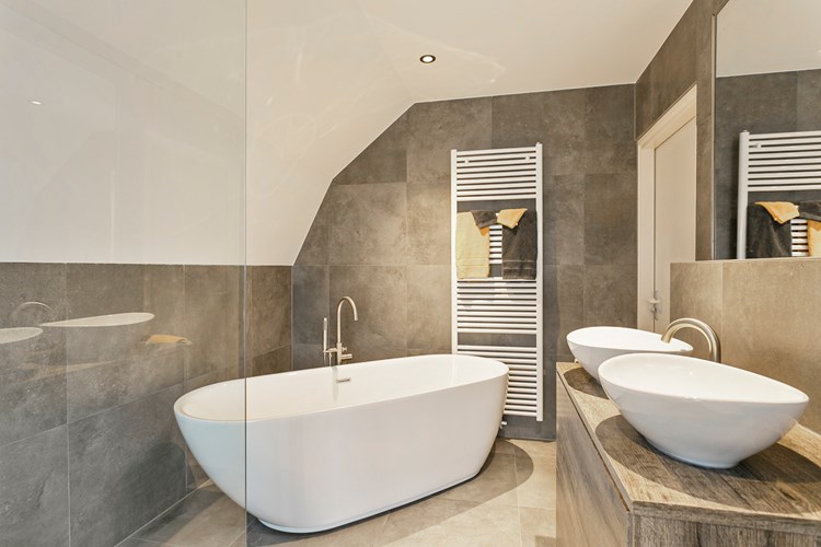 Een luxe badkamer (2021) met een tegelvloer, grotendeels betegelde wanden en een stucwerk plafond met LED inbouwspots. Voorzien van een badmeubel met twee waskommen en een grote spiegel, een kunststof ligbad met thermostaatkraan. 
