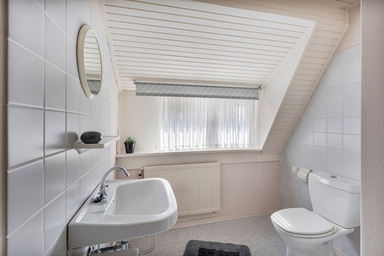 Een ruime toiletruimte met een vinylvloer, betegelde wanden en een schroten plafond. Met een duoblok, een wastafel en een radiator. Daglicht en natuurlijke ventilatie via een dakkapel met houten kozijn met enkele beglazing.