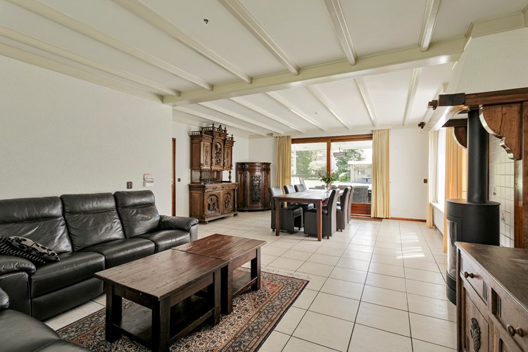 Royale woonkamer met een lichte tegelvloer met vloerverwarming, granol- en schoonmetselwerk wanden en een licht sierbalken plafond. 