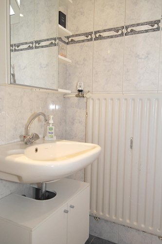 Volledig licht betegelde badkamer met een kunststof panelen plafond met inbouwspots. Met een wastafel met onderkastje, een spiegelkast en een radiator. 