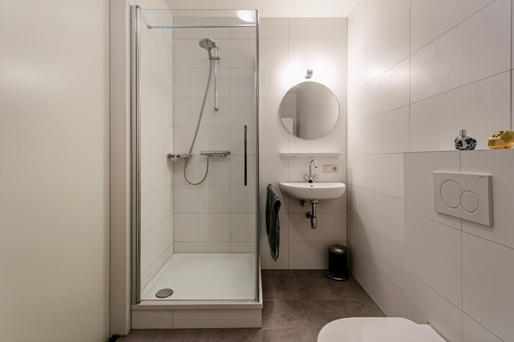 De volledig licht betegelde badkamer is voorzien van een douchecabine met glazen deur en thermostaatkraan, een wandcloset en een wastafel met planchet en spiegel. 