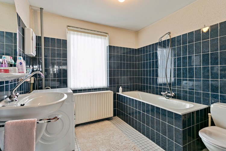 Ruime badkamer met een lichte tegelvloer, gedeeltelijke betegelde wanden en een stucwerk plafond. Met een ligbad en natuurlijke ventilatie middels een hardhouten raamkozijn met enkele beglazing. 