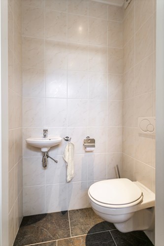 Het moderne toilet is voorzien van een antraciet tegelvloer en volledig licht betegelde wanden. Met een wandcloset, een fonteintje en mechanische ventilatie.