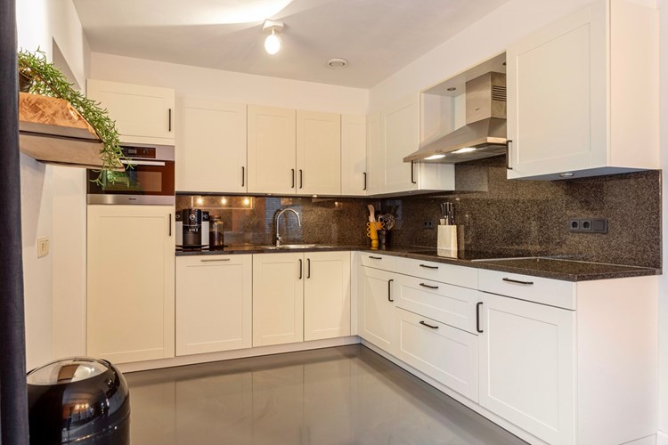 De moderne, lichte keukeninrichting in een hoekopstelling is voorzien van een granieten aanrechtblad en -achterwand, een RVS spoelbak met restbakje, een inductiekookplaat, een RVS afzuigkap, een vaatwasser, een koelkast en een combimagnetron. Vanuit de keuken met een deur naar de bijkeuken.
