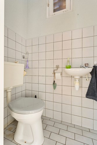 Toilet met een lichte tegelvloer, gedeeltelijk licht betegelde wanden en een spuitwerk plafond. Voorzien van een duoblok, een fonteintje en een raampje. 
