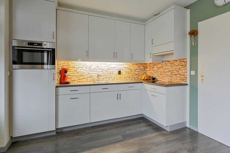 Een lichte, moderne keukeninrichting in een hoekopstelling voorzien van een RVS spoelbak, een keramische kookplaat, een afzuigkap, een koelkast en een RVS combimagnetron. Onder de trapopgang bevindt zich een praktische voorraadkast. 