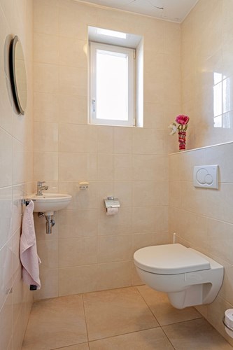 Een volledig licht betegeld toilet met een wandcloset met opzetplateau en een hoekfonteintje. Natuurlijke ventilatie middels een kunststof raamkozijn (draai-/kiep) met dubbele beglazing. 
