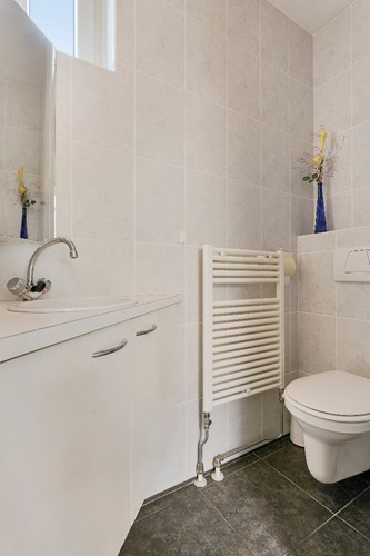 Bij de wasruimte ligt ook het volledig, licht betegelde toilet. Met een wandcloset met opzetplateau en een meubel met fonteintje. Natuurlijke ventilatie middels een kunststof raampje.