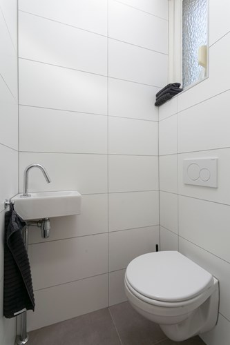 Modern toilet met een antraciet tegelvloer en volledig licht betegelde wanden. Met een wandcloset en een hoekfonteintje. Natuurlijke ventilatie middels een raampje met dubbel bewerkt glas en een rolluik. 