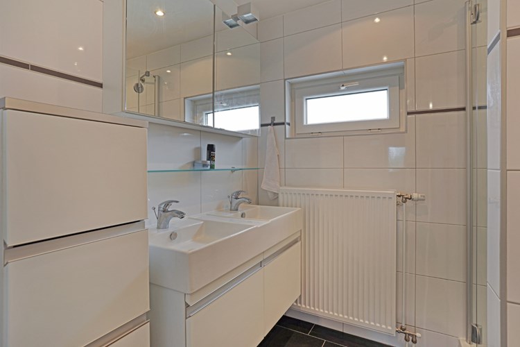 Een moderne badkamer met een antraciet tegelvloer met vloerverwarming, volledig licht betegelde wanden en een stucwerk plafond. Met een badmeubel met dubbele wastafel en een spiegelkast. 