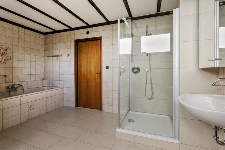 Royale badkamer met een tegelvloer, volledig betegelde wanden en een sierbalken plafond. Met een ruime douchecabine met in de wand geïntegreerde kranen en een ligbad met een opzetplateau. 