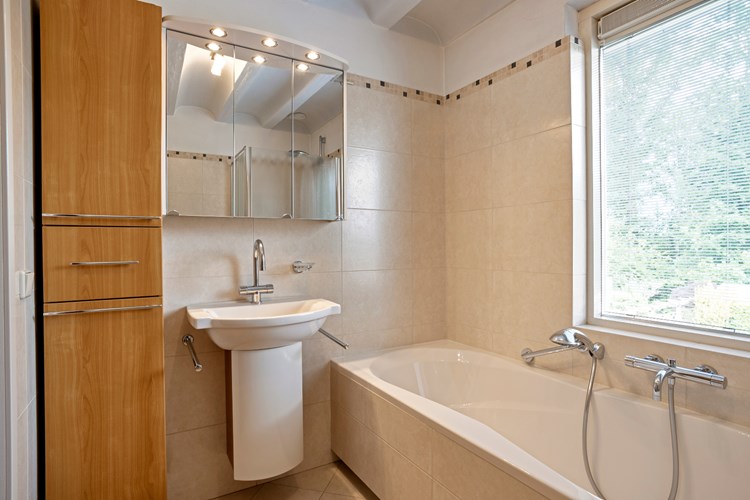 Een moderne badkamer met een lichte tegelvloer, grotendeels betegelde wanden en een gewelfd plafond. Met een kunststof ligbad met thermostaatkraan en een badmeubel met wastafel en een spiegel met verlichting. 
