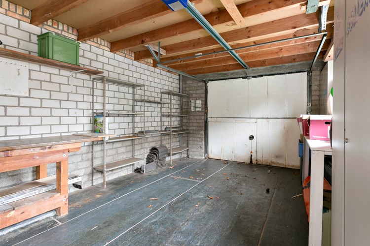 De in spouw gemetselde garage is voorzien van een plat (bitumen) dak. Voorzien van elektra en aan de voorzijde een op afstand bedienbare stalen kantelpoort. Daglicht via een hardhouten raamkozijn en een deur in de zijgevel aan de tuinzijde.