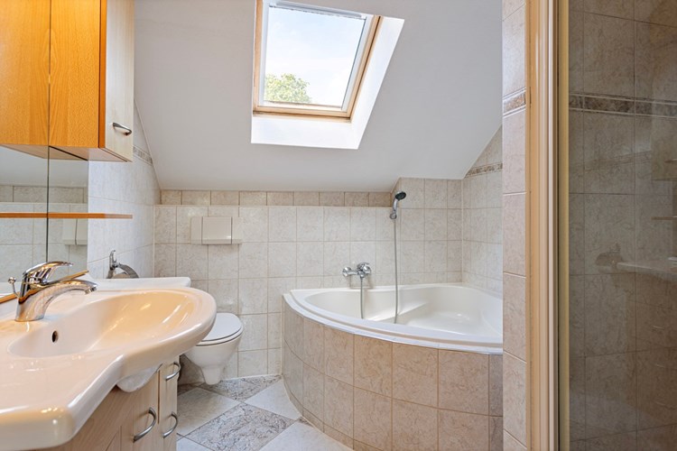 Ruime badkamer met een lichte tegelvloer, volledig licht betegelde wanden en een spuitwerk plafond. Voorzien van een hoek-/zit ligbad, een wandcloset en een badmeubel met wastafel en een spiegel met inbouwspots. 