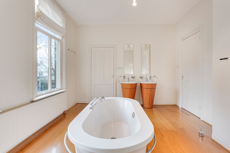 Een ruime luxe, design badkamer met een parketvloer. Voorzien van een vrijstaand ligbad en een tweetal design wastafels met een spiegel. 