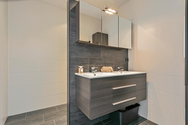 Een moderne badkamer met een antraciet tegelvloer met vloerverwarming, volledig licht betegelde wanden en een kunststof panelen plafond met inbouwspots. Met een badmeubel met dubbele wastafel met een spiegelkast. 