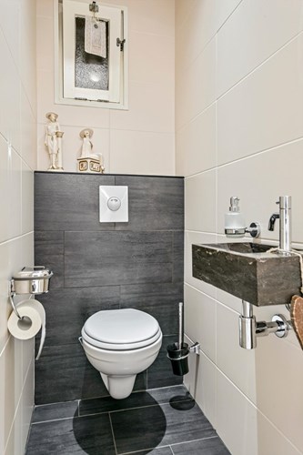 Volledig betegeld toilet met een antraciet tegelvloer met vloerverwarming, volledig licht betegelde wanden en een kunststof panelen plafond met inbouwspots. Met een wandcloset met een opzetplateau, een hardstenen fonteintje en een raampje. 