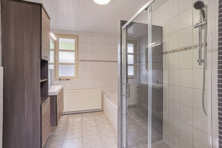 Ruime badkamer met een lichte tegelvloer, volledig licht betegelde wanden en een kunststof schroten plafond. Met een ligbad en een inloopdouche met een schuifwand. 