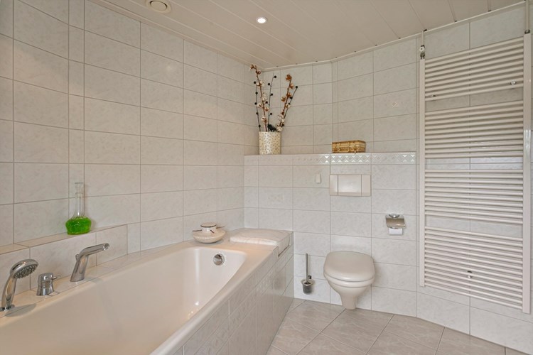 Een volledig licht betegelde badkamer en een MDF panelen plafond met inbouwspots. Voorzien van een kunststof ligbad met geïntegreerde kranen en een wandcloset met opzetplateau. 