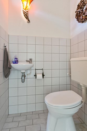 Toilet met een lichte tegelvloer, gedeeltelijk licht betegelde wanden en een stucwerk plafond. Met een toilet en een fonteintje. Natuurlijke ventilatie middels een raampje. 