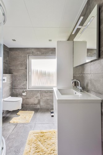 Een moderne badkamer (2021) met een tegelvloer, volledig betegelde wanden en een panelen plafond met inbouwspots. Met een wandcloset met een opzetplateau en een badmeubel met een wastafel en een grote spiegel met verlichting.  