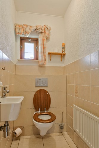 Toilet met een lichte tegelvloer, gedeeltelijk betegelde wanden en een stucwerk plafond. Met een wandcloset met een opzetplateau, een fonteintje en een raampje. 