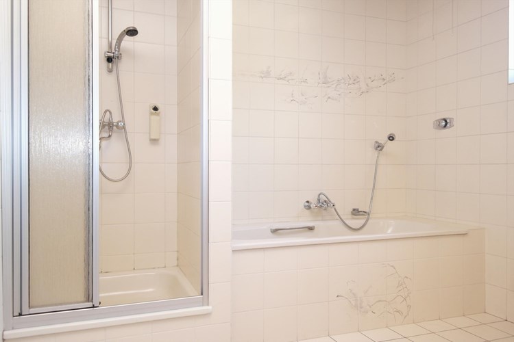 Badkamer met een lichte tegelvloer, volledig licht betegelde wanden en een schroten plafond. Voorzien van een douchecabine en een ligbad. 