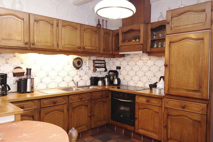 Vanuit de woonkamer zicht op de open keuken met een eiken keukeninrichting voorzien van een eiken aanrechtblad, twee RVS spoelbakken, een inductiekookplaat, een afzuigkap, een elektrische oven, een koelkast en een vaatwasser.