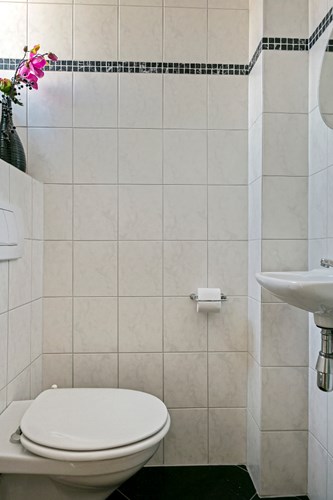 Toilet met een antraciet tegelvloer, volledig licht betegelde wanden en een MDF plafond. Met een wandcloset met opzetplateau en een fonteintje. Natuurlijke ventilatie middels een hardhouten raamkozijn met dubbel gematteerd glas. 