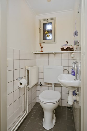 Toilet met een tegelvloer, gedeeltelijk licht betegelde wanden en een stucwerk plafond. Met een duoblok en een fonteintje. Natuurlijke ventilatie middels een raampje. 