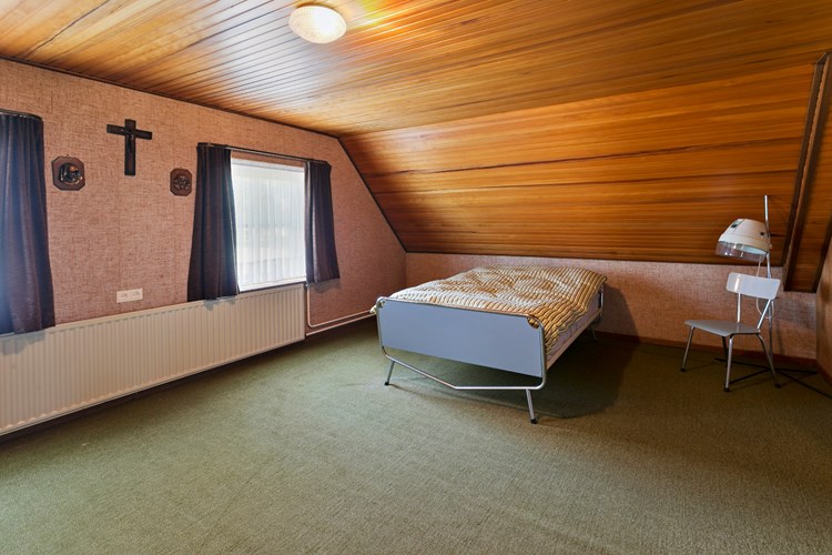 Ruime slaapkamer gelegen aan de linkerzijde van de woning, met vloerbedekking, behangen wanden en een schroten plafond. 