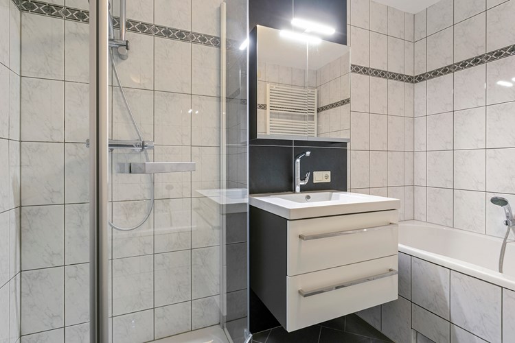 Badkamer met een tegelvloer met vloerverwarming, volledig licht betegelde wanden en een spuitwerk plafond. Met een kunststof ligbad en een badmeubel met vaste wastafel en een spiegelkast. 