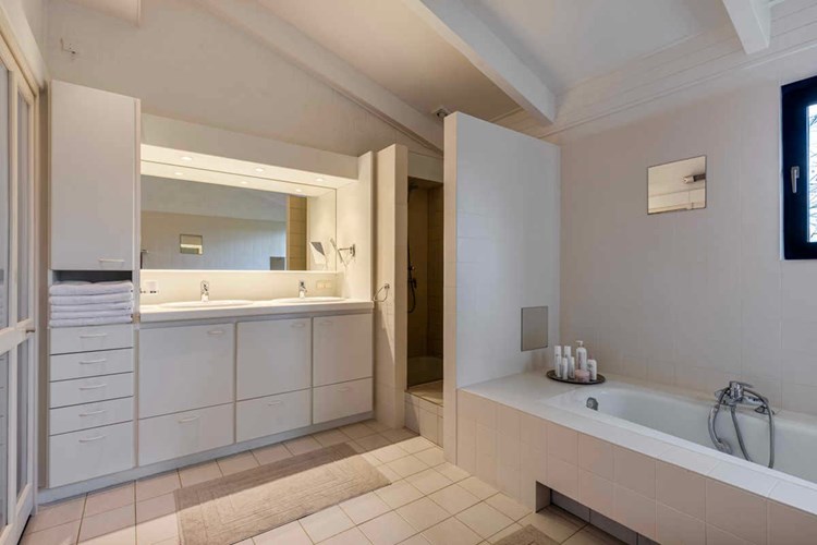 De ruime, volledig licht betegelde badkamer met een oplopend plafond is voorzien van een badmeubel met dubbele wastafel en een grote spiegel. 