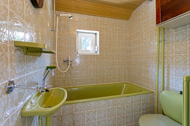 Badkamer met een tegelvloer, volledig betegelde wanden en een kunststof schroten plafond. Met een ligbad met douchemogelijkheid, een 2e toilet en een fonteintje. Natuurlijke ventilatie middels een kunststof raampje (draai-/kiep) met een rolluik. 