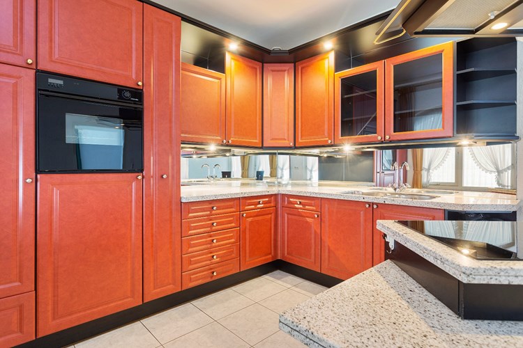 Een keukeninrichting in een hoekopstelling met een granieten aanrechtblad en een spiegel achterwand. Voorzien van een dubbele RVS spoelbak, een koelkast, een diepvries (4-laden) en een elektrische oven (defect). Verder een kookeiland met een inductiekookplaat en een schouw afzuigkap. 