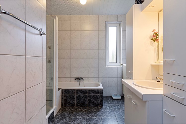 Badkamer met een tegelvloer met vloerverwarming, volledig licht betegelde wanden en een kunststof panelen plafond. Met  een stalen ligbad met een opzetplateau. 