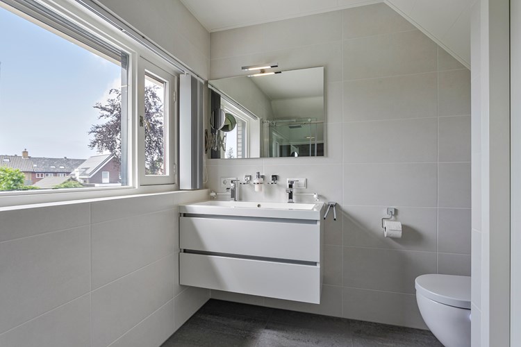 Moderne badkamer (2015) met een antraciet tegelvloer met elektrische vloerverwarming, licht betegelde wanden en een panelen plafondafwerking. Voorzien van een wandcloset (Sanibroyeur) en een badmeubel met vaste wastafel, een tweetal kranen en een spiegel met verlichting. 
