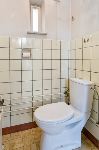Toilet met een tegelvloer, gedeeltelijk betegelde wanden en een sierbalken plafond. Voorzien van een duoblok en een raampje. 