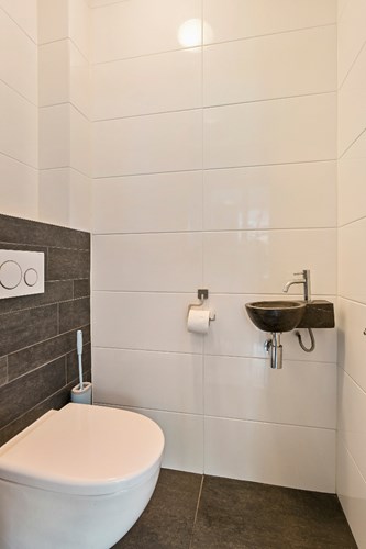 Een modern toilet (2016) voorzien van een antraciet tegelvloer, volledig licht betegelde wanden en een stucwerk plafond. Met een wandcloset met een opzetplateau en een hardstenen fonteintje. Mechanische ventilatie is aanwezig. 