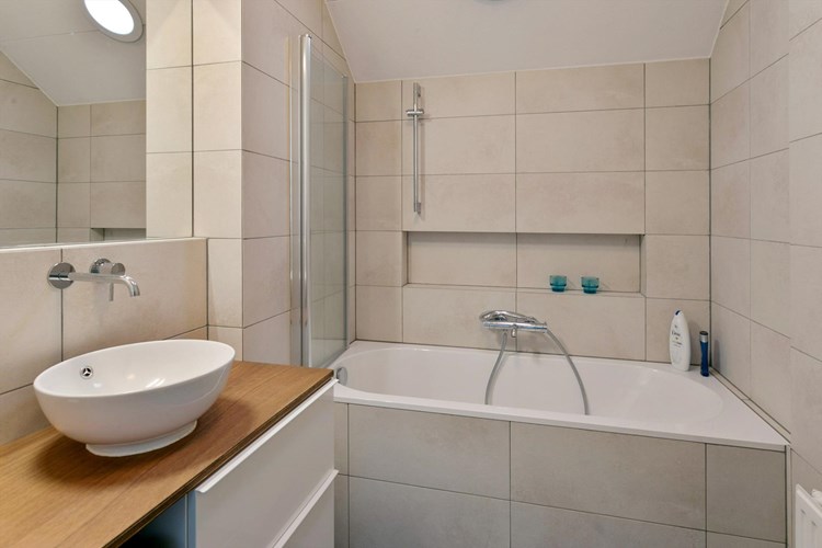 Moderne badkamer met een tegelvloer, volledig licht betegelde wanden en een spuitwerk plafond. Voorzien van een badmeubel met waskom, geïntegreerde kraan en een grote spiegel en een kunststof ligbad met douche garnituur en een glazen spatwand. Daglicht middels een solatube. 
